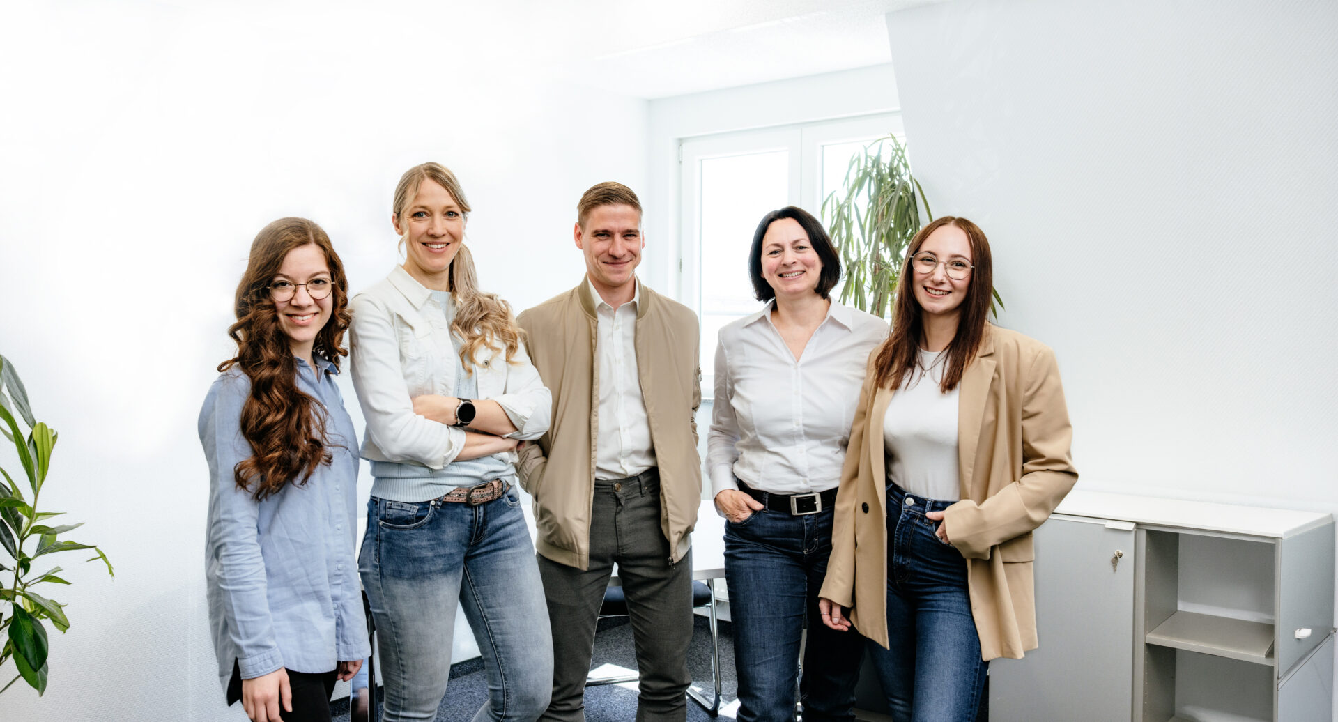 Fünf lächelnde Personen in legerer Kleidung stehen in einem hellen Büro mit Pflanzen und Regalen